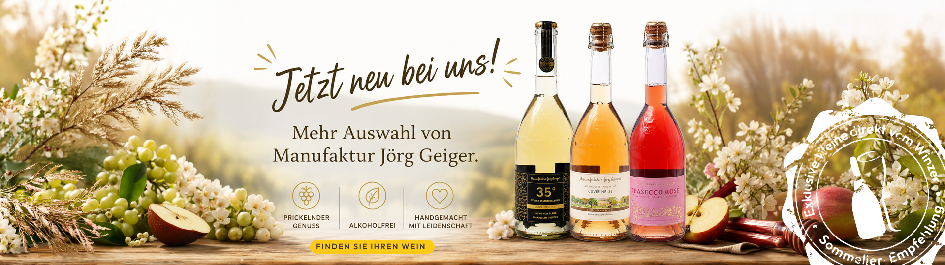 Drei Weinflaschen unterschiedlicher Form und Farbe stehen auf einem Holztisch vor einem hellen Hintergrund mit Weintrauben und Getreidehalmen, darüber der Text 'Jetzt neu bei uns! Mehr Auswahl von Manufaktur Jörg Geiger.'